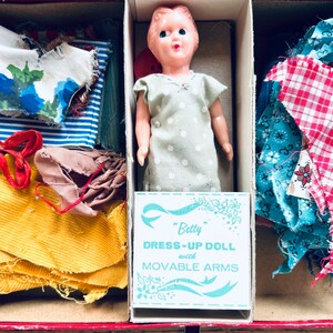 May include: A vintage doll dress-up kit featuring a plastic doll, fabric scraps, and a label that reads "Betty Dress-Up Doll with Movable Arms".