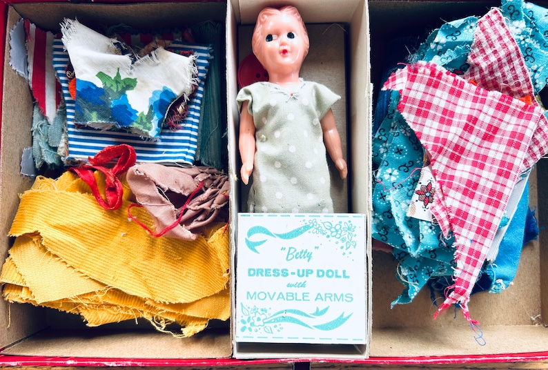 May include: A vintage doll dress-up kit featuring a plastic doll, fabric scraps, and a label that reads "Betty Dress-Up Doll with Movable Arms".