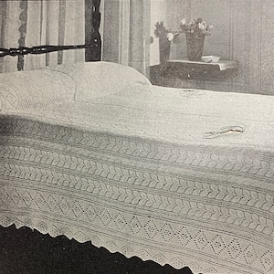 May include: A white crocheted bedspread with a diamond pattern and a scalloped edge. The bedspread is on a wooden bed frame with a white pillow.