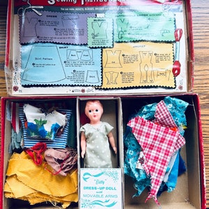 May include: A vintage doll dress-up kit featuring a doll, fabric scraps, and instructions for making clothes. The kit includes a doll with movable arms, a yellow fabric, a blue and white striped fabric, a red and white checked fabric, a blue fabric with white flowers, and a brown fabric. The box lid has instructions for making a dress, blouse, skirt, jacket, and buttonhole stitch.