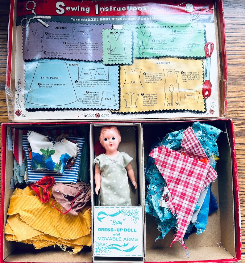 May include: A vintage doll dress-up kit featuring a doll, fabric scraps, and instructions for making clothes. The kit includes a doll with movable arms, a yellow fabric, a blue and white striped fabric, a red and white checked fabric, a blue fabric with white flowers, and a brown fabric. The box lid has instructions for making a dress, blouse, skirt, jacket, and buttonhole stitch.