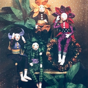 May include: A collection of handmade fabric dolls with flower-shaped heads and whimsical outfits. The dolls feature painted faces and are dressed in various colors, including orange, purple, green, and blue. They are posed among greenery and a decorative wreath.
