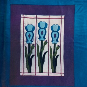 Può includere: Un'opera d'arte incorniciata con tre fiori blu con steli e foglie verdi su uno sfondo bianco. La cornice è viola con un bordo bianco.