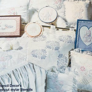 May include: An assortment of white and pastel-toned embroidered cushions, framed art, and stencils. The artwork depicts a duck scene, a teddy bear, and floral patterns. The text "Stencil Decor's pre-cut mylar Stencils" is visible.