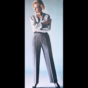 May include: A woman wearing a white blouse and gray high-waisted pants. She is standing with her arms crossed and looking to the side.