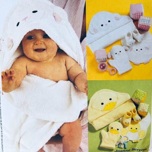 May include: A baby wrapped in a white hooded towel with a sheep face design. The image also shows a matching set of baby bath accessories, including a washcloth, booties, and a rattle. A second set features a yellow duck design.