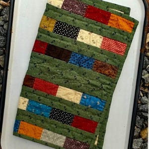 May include: A small, rectangular quilt with a patchwork design. The quilt features rows of colorful fabric rectangles in shades of red, orange, blue, brown, and cream, set against a green background. The quilt is folded over.