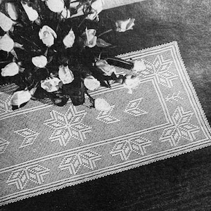 May include: A white crocheted table runner with a geometric pattern. The runner is laid on a dark surface with a bouquet of white flowers on top.