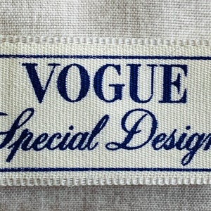 May include: A rectangular woven label with the words "VOGUE Special Design" in navy blue. The text is enclosed in a navy blue rectangular border. The label is on a light beige fabric.