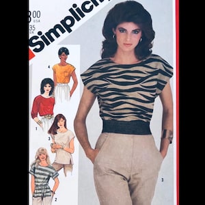 Può includere: Una donna indossa un top beige e nero a righe zebrate e pantaloni color cachi. Il top ha maniche corte e una fascia in vita nera. L'immagine è tratta da un modello di cucito Simplicity, con il testo "Simplicity" e i numeri del modello visibili.