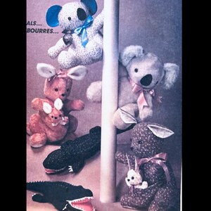 Puede incluir: Una colección de animales de peluche hechos a mano, incluyendo koalas, canguros y caimanes. Los koalas tienen lazos azules, los canguros tienen lazos rosas y los caimanes son negros con bocas rojas. Los animales de peluche están hechos de tela suave.