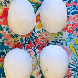 May include: Four white, egg-shaped bath bombs are arranged on a colorful floral patterned fabric. The bath bombs have a textured surface and are a solid white color. The fabric background features red, pink, blue, and green floral designs.