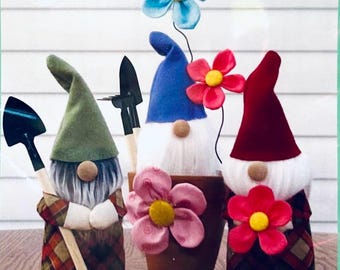 May include: Three garden gnomes with white beards, holding miniature gardening tools. One gnome wears a green hat, another a blue hat, and the third a red hat. Each gnome has a plaid patterned body and is decorated with colorful fabric flowers.