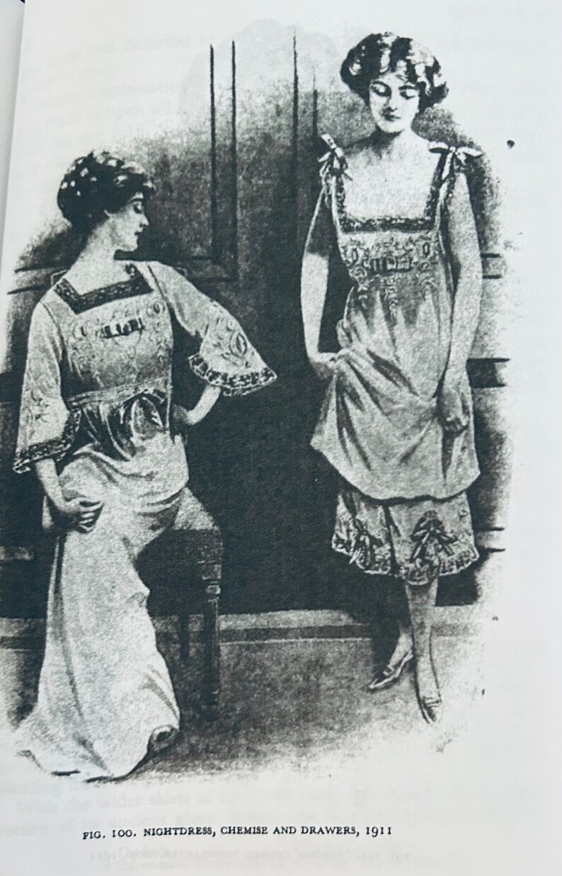 May include: Black and white photo of two women wearing nightgowns. The woman on the left is sitting on a chair and the woman on the right is standing. The caption below the image reads "Fig. 100. Nightdress, Chemise and Drawers, 1911."