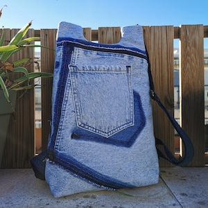 Op de afbeelding: Een lichtblauwe denim rugzak met donkerblauwe details, waaronder een zak en afwerking. De tas heeft een zwarte rits en een donkerblauwe band. Het ontwerp bevat de achterzak van een spijkerbroek.