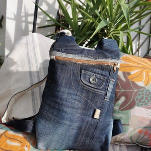 Puede incluir: Una mochila vaquera con cierre de cremallera y un bolsillo delantero. La bolsa está hecha de vaqueros azules reciclados y tiene una correa de color claro. La mochila está sobre un cojín con estampado floral.