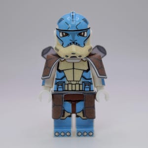 May include: A blue and tan Lego minifigure of a clone trooper with a helmet, armor, and brown shoulder pads. The figure has white hands and is standing upright. The figure is a collectible toy.