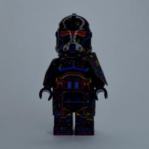 May include: A black Lego minifigure with a helmet and armor, decorated with colorful patterns. The figure has a red and yellow design on the helmet and chest, with blue and yellow accents. The figure is standing upright.