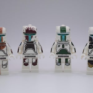 Puede incluir: Cuatro minifiguras de Lego con armadura blanca y varios acentos de color. Cada figura tiene un casco, una armadura para el pecho y detalles en las piernas. Los esquemas de color incluyen marrón, verde y negro, con viseras turquesas. Las figuras están de pie.