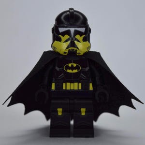 May include: A black and yellow Lego minifigure with a Batman theme. The minifigure has a black helmet with yellow accents, a black cape, and a chest emblem. The figure's design incorporates elements from both Batman and Star Wars.