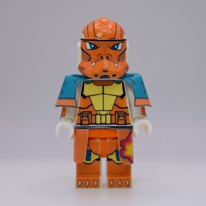 May include: A Lego minifigure of a clone trooper in orange and yellow armor with a blue cape. The helmet has a detailed design with blue eyes. The figure has white hands and is standing upright.