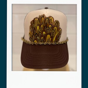 May include: A brown trucker hat with a tan front panel adorned with a cluster of yellow and brown feathers. A gold chain encircles the base of the feather design, and the brim is a solid brown color.