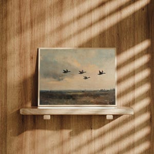 K&ouml;nnte beinhalten: Ein gerahmtes Gem&auml;lde, das eine Landschaft mit f&uuml;nf V&ouml;geln zeigt, die in Formation vor einem bew&ouml;lkten Himmel fliegen. Das Gem&auml;lde befindet sich auf einem Holzregal an einer Holzvert&auml;felung.
