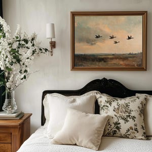 K&ouml;nnte beinhalten: Ein wei&szlig;es Bett mit einer wei&szlig;en gesteppten Bettdecke und einem schwarzen Kopfteil. Auf dem Bett liegen mehrere Kissen, darunter ein cremefarbenes Kissen, ein wei&szlig;es Kissen und ein Kissen mit Blumenmuster. Ein gerahmtes Gem&auml;lde von V&ouml;geln, die &uuml;ber eine Landschaft fliegen, h&auml;ngt an der Wand hinter dem Bett.
