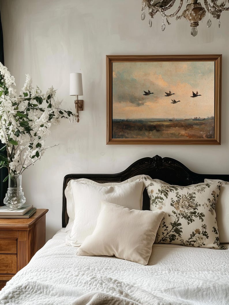 K&ouml;nnte beinhalten: Ein wei&szlig;es Bett mit einer wei&szlig;en gesteppten Bettdecke und einem schwarzen Kopfteil. Auf dem Bett liegen mehrere Kissen, darunter ein cremefarbenes Kissen, ein wei&szlig;es Kissen und ein Kissen mit Blumenmuster. Ein gerahmtes Gem&auml;lde von V&ouml;geln, die &uuml;ber eine Landschaft fliegen, h&auml;ngt an der Wand hinter dem Bett.