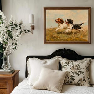 May include: A white bed with a white duvet cover and a black headboard. There are three pillows on the bed, one with a floral pattern and two plain white pillows. A framed painting of two dogs running through a field hangs on the wall above the bed.