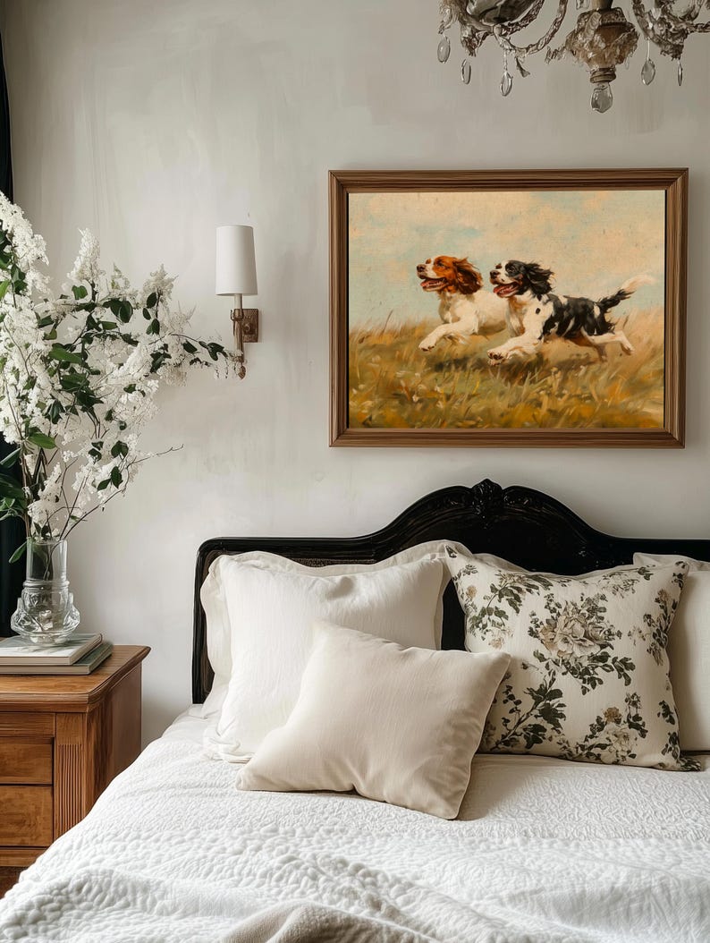 May include: A white bed with a white duvet cover and a black headboard. There are three pillows on the bed, one with a floral pattern and two plain white pillows. A framed painting of two dogs running through a field hangs on the wall above the bed.
