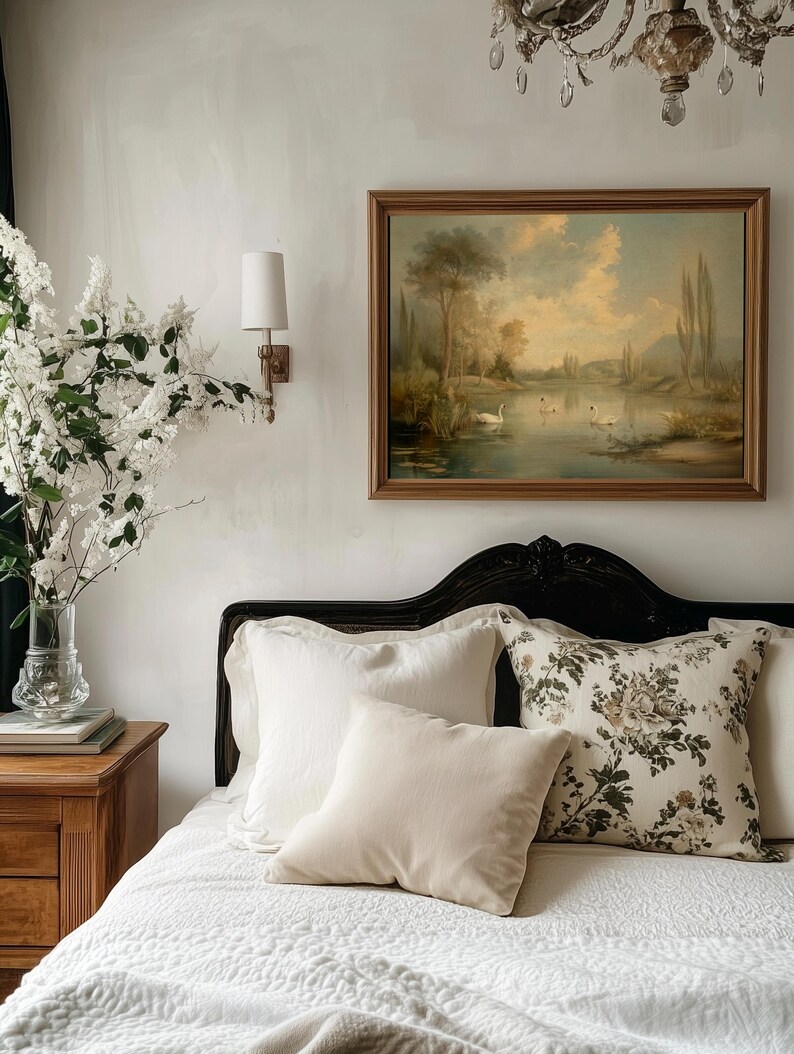 K&ouml;nnte beinhalten: Ein wei&szlig;es Bett mit einem schwarzen Kopfteil und wei&szlig;en Bettw&auml;sche. Auf dem Bett liegen mehrere Kissen, darunter ein Kissen mit Blumenmuster und ein schlichtes wei&szlig;es Kissen. &Uuml;ber dem Bett h&auml;ngt ein Gem&auml;lde mit einer Seenszene.