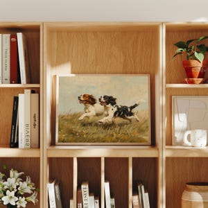 May include: A vintage painting of two dogs running through a field. The dogs are a brown and white Springer Spaniel and a black and white Springer Spaniel. The painting is framed in a light wood frame.