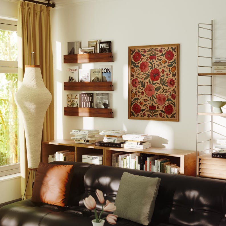 May include: A living room scene featuring a black leather sofa with orange and green pillows, a wooden bookshelf, and a framed floral artwork. Books are displayed on shelves and a tall, cream-colored lamp stands near a window with beige curtains.