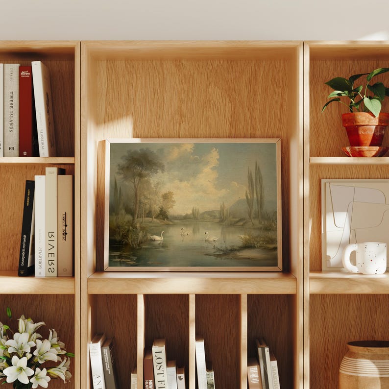 K&ouml;nnte beinhalten: Ein gerahmtes Landschaftsbild eines Sees mit B&auml;umen und Schw&auml;nen in einem B&uuml;cherregal aus Holz mit anderen B&uuml;chern und Pflanzen.