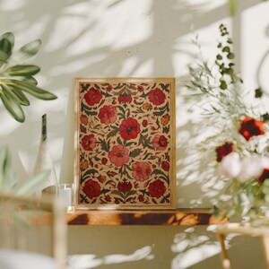 May include: A framed floral print featuring a repeating pattern of red flowers and green leaves on a beige background. The wooden frame complements the artwork, creating a decorative piece. The print is displayed on a wooden shelf, with plants and flowers in the background.