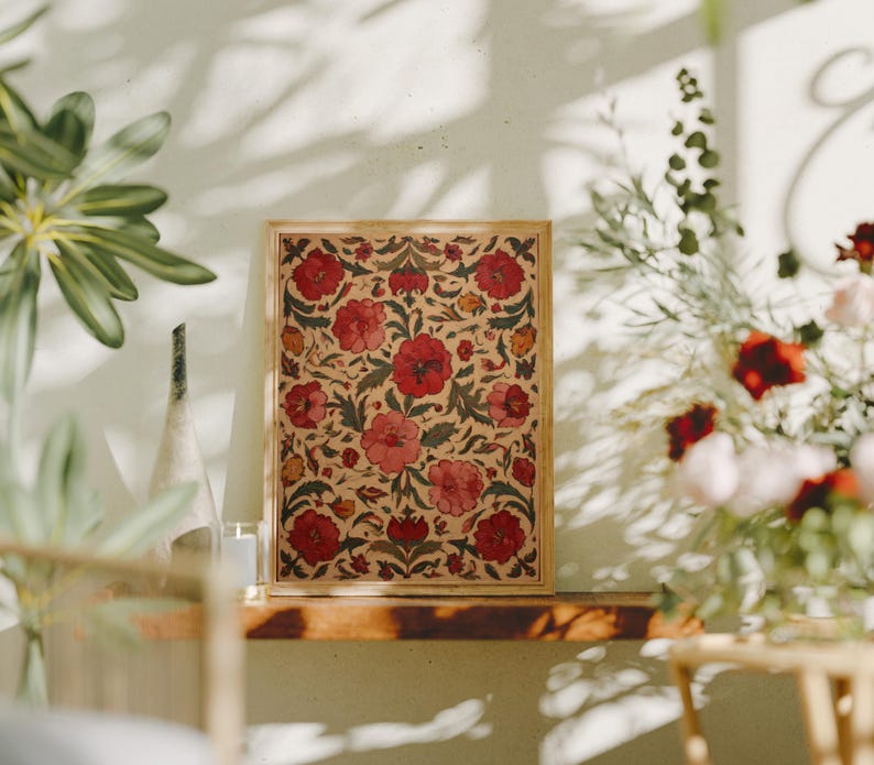 May include: A framed floral print featuring a repeating pattern of red flowers and green leaves on a beige background. The wooden frame complements the artwork, creating a decorative piece. The print is displayed on a wooden shelf, with plants and flowers in the background.