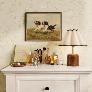 May include: A white dresser with a drawer and a white lamp with a cream-colored shade and a green trim. On the dresser are various items, including a book titled "L'Amour du Cinema", a glass bottle of perfume, a small glass jar with a lid, a wooden tray with a white marble, and a white candle with the word "White" printed on it.