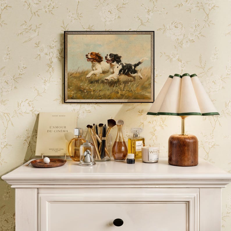 May include: A white dresser with a drawer and a white lamp with a cream-colored shade and a green trim. On the dresser are various items, including a book titled "L'Amour du Cinema", a glass bottle of perfume, a small glass jar with a lid, a wooden tray with a white marble, and a white candle with the word "White" printed on it.