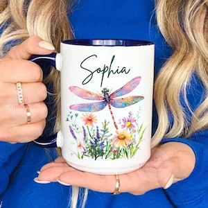 May include: White ceramic mug with a navy blue interior and handle. The mug features a colourful dragonfly design with the name "Sophia" in script. Floral accents include daisies and lavender. The mug is 425ml.