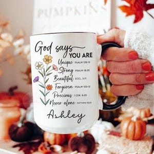 May include: White ceramic mug with a black handle and rim, inscribed with "God says YOU ARE" and inspirational words, decorated with floral designs. The mug is personalized with the name "Ashley".
