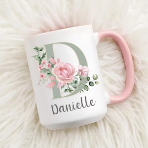 May include: A white ceramic mug with a 15 oz capacity, featuring a large sage green letter D decorated with pink roses and green leaves. The name Danielle is printed in a dark gray script font below the letter. The mug has a light pink handle.
