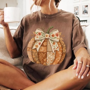 May include: A brown t-shirt featuring a patchwork pumpkin design with a floral bow. The pumpkin is composed of various orange and brown patterned fabrics. The shirt is a relaxed fit.
