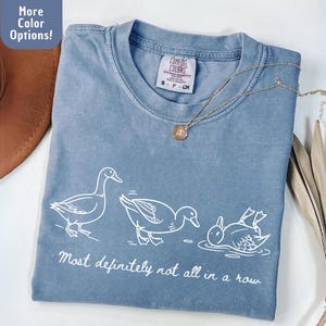 May include: A blue Comfort Colors t-shirt with a white graphic of three ducks in different poses and the text "Most definitely not all in a row." A gold necklace is draped across the shirt.