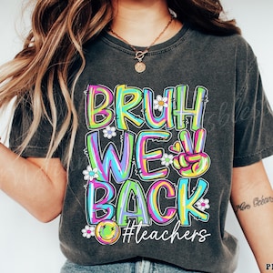May include: Dark gray t-shirt with the words "BRUH WE BACK" in colorful, bubble letters. The design includes a peace sign, daisies, and a smiley face. The hashtag "#teachers" is also included in the design.