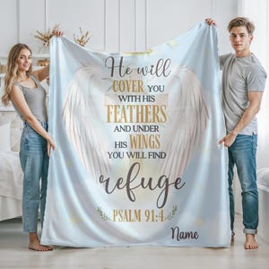 May include: A light blue blanket with a design featuring angel wings and the text "He will cover you with his feathers and under his wings you will find refuge Psalm 91:4 Name". The blanket is held by two people.