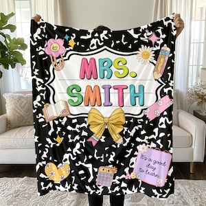 May include: A black and white patterned blanket with the name "MRS. SMITH" in colorful letters. The blanket features school-themed illustrations like a pencil, book, and calculator. A large yellow bow and the phrase "It's a good day to learn" are also displayed.