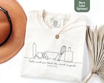May include: A cream-colored t-shirt featuring a black line art graphic of kitchen tools including a rolling pin, mug, bowl, whisk, and cutting board. Below the graphic is the text Taste and see that the Lord is good. PSALM 34:8.