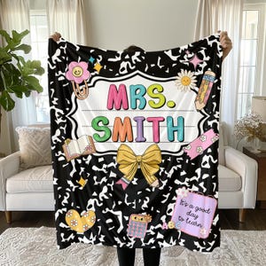 May include: A black and white patterned blanket with the name "MRS. SMITH" in colorful letters. The blanket features school-themed illustrations like a pencil, book, and calculator. A large yellow bow and the phrase "It's a good day to learn" are also displayed.