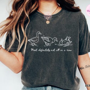 May include: Dark gray t-shirt with a white line drawing of three ducks, one upside down. The text "Most definitely not all in a row" is below the ducks. The shirt is a casual style.